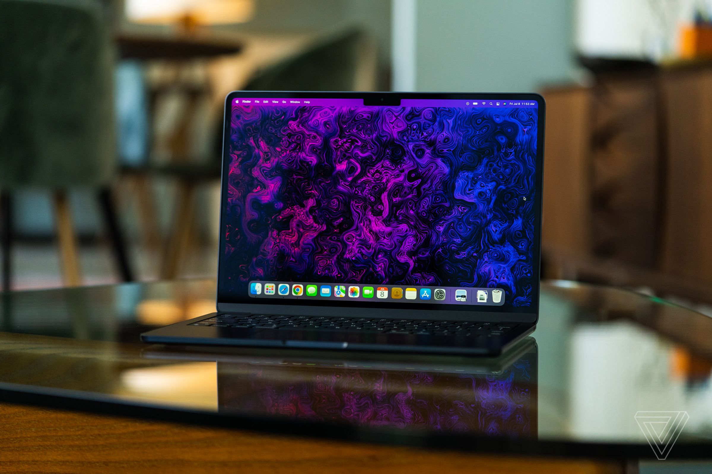 The M2 MacBook Air is opened, facing the camera. Its display is on, showcasing a psychedelic purple and black wallpaper created by The Verge's art and illustration team.