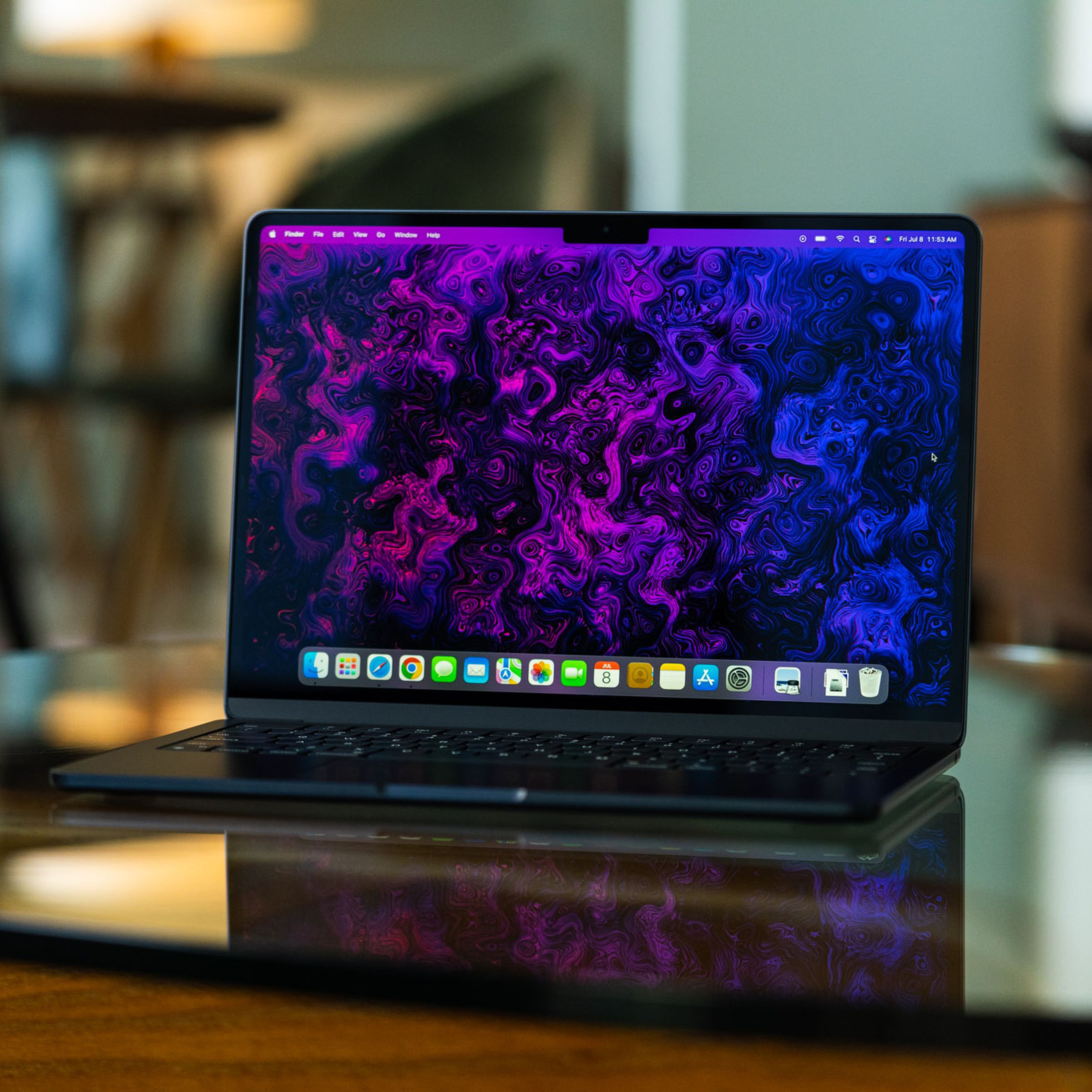 The M2 MacBook Air is opened, facing the camera. Its display is on, showcasing a psychedelic purple and black wallpaper created by The Verge's art and illustration team.