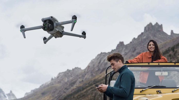 DJI Air 3 in flight by a mountain, with person looking at controller alongside yellow four wheel drive vehicle