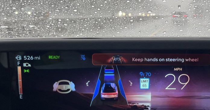 Close-up of dashboard with warning “keep hands on steering wheel” and rain on windshield.