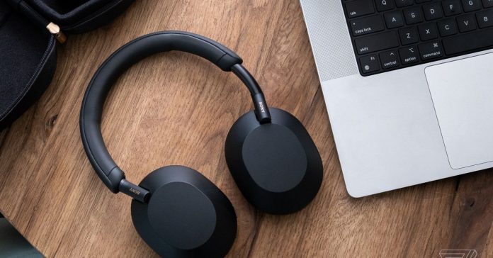 A pair of Sony WH-1000XM5 headphones, in black, resting flat on a wood table beside a laptop.