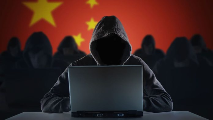 A group of 7 hackers, 6 slightly blurred in the background and one in the foreground, all wearing black with hoods pulled up over their heads. You cannot see their faces. The hacker in the foreground sits with an open laptop in front of them. The background, behind the hackers, is a Chinese flag