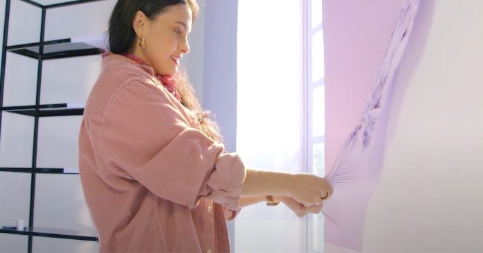 A person peels a thin layer of purple paint off of a wall.